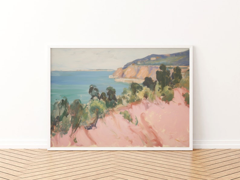 May include: A framed landscape painting featuring a coastal scene with a pink foreground, green trees, and a blue sea meeting a distant cliffside under a pale sky. The artwork is enclosed in a simple white frame, displaying soft brushstrokes.