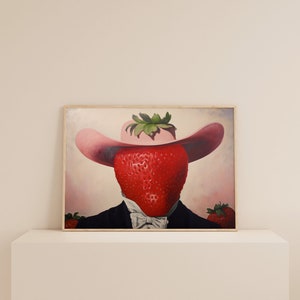 Peut inclure: Une impression d'art encadr&eacute;e repr&eacute;sentant une illustration surr&eacute;aliste d'une grosse fraise rouge portant un chapeau rose &agrave; larges bords et une veste de costume noire avec un n&oelig;ud papillon blanc. Deux petites fraises sont visibles en bas.