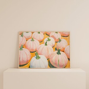 Op de afbeelding: Een rechthoekig ingelijst schilderij met een verzameling lichtroze en witte pompoenen met groene stelen, gerangschikt op een felgeel oppervlak. Het kunstwerk toont meerdere pompoenen van verschillende groottes, weergegeven met zichtbare penseelstreken en zachte highlights.