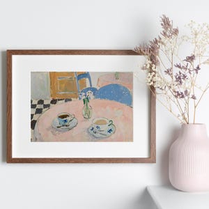 May include: A framed art print depicting a cafe scene with two teacups on a pink table, a small vase of flowers, and a black and white checkered floor. Next to the frame sits a light pink ribbed ceramic vase containing dried floral stems.