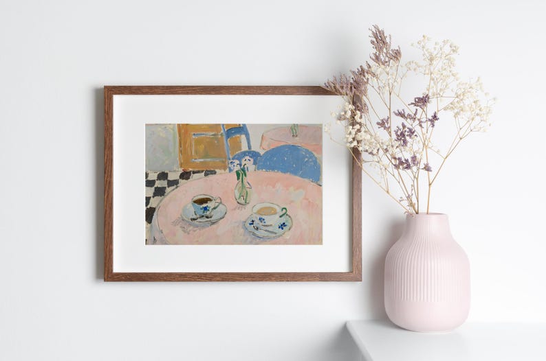 May include: A framed art print depicting a cafe scene with two teacups on a pink table, a small vase of flowers, and a black and white checkered floor. Next to the frame sits a light pink ribbed ceramic vase containing dried floral stems.