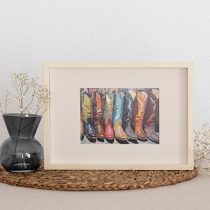 May include: A framed print featuring a row of colorful cowboy boots in shades of yellow, orange, red, blue, black, and brown. The artwork is displayed in a light-colored wooden frame, showcasing a detailed illustration of various boot styles.