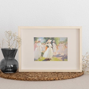 May include: A framed art print featuring a painting of figures in white dresses and hats in a sunny outdoor setting. The artwork is in a light wooden frame, displayed on a woven mat, with a dark gray vase and dried flowers nearby.