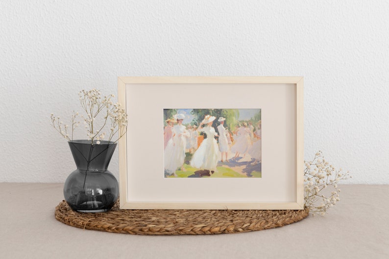 May include: A framed art print featuring a painting of figures in white dresses and hats in a sunny outdoor setting. The artwork is in a light wooden frame, displayed on a woven mat, with a dark gray vase and dried flowers nearby.