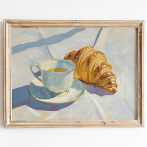 May include: A painting of a white teacup and saucer with a golden yellow croissant on a white tablecloth. The painting is framed in a light wood frame.