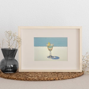 May include: A framed painting of a cocktail with a lime wedge in a martini glass. The painting is on a light blue background with a white stripe.