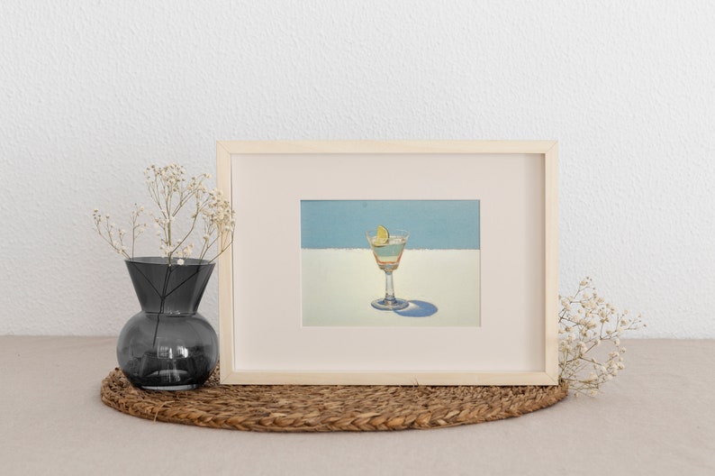 May include: A framed painting of a cocktail with a lime wedge in a martini glass. The painting is on a light blue background with a white stripe.
