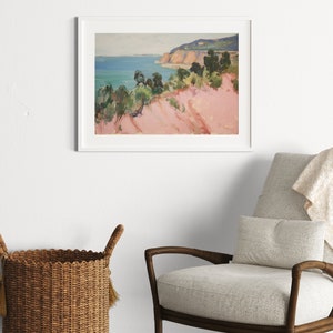 May include: A framed landscape painting depicting a coastal scene with a pinkish-toned foreground, green trees, and a blue ocean under a pale sky. The artwork is enclosed in a simple white frame and is displayed on a plain wall above a chair.