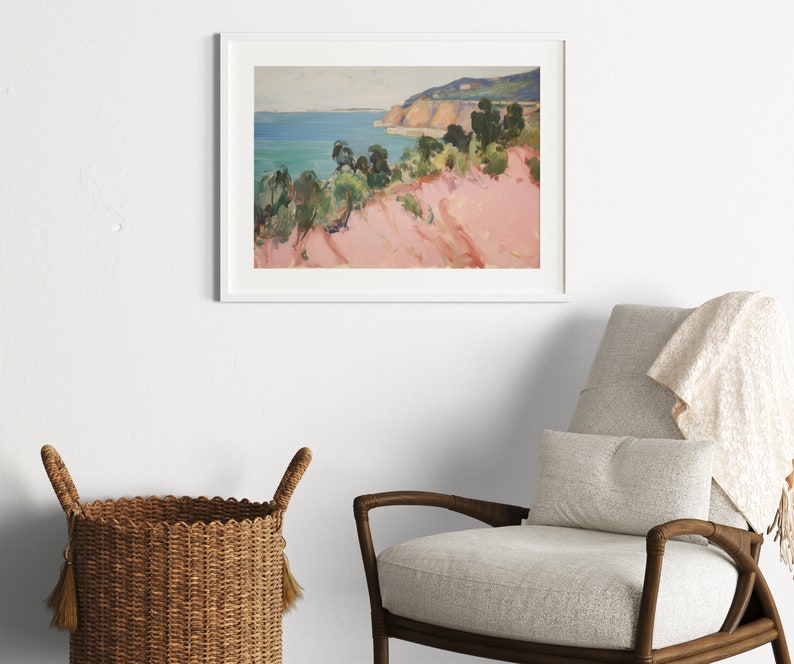 May include: A framed landscape painting depicting a coastal scene with a pinkish-toned foreground, green trees, and a blue ocean under a pale sky. The artwork is enclosed in a simple white frame and is displayed on a plain wall above a chair.