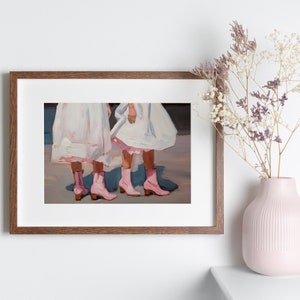 May include: Painting of two people wearing white dresses and pink cowboy boots. The painting is framed in a brown wood frame.