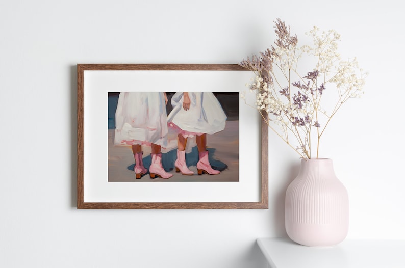 May include: Painting of two people wearing white dresses and pink cowboy boots. The painting is framed in a brown wood frame.