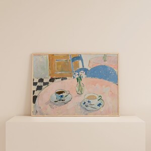 May include: A framed rectangular art print depicting a painted interior scene with a round pink table, two teacups on saucers, and a small vase with flowers. The background features a black and white checkered floor, a blue chair, and a wooden cabinet.