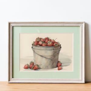 May include: A framed watercolor painting of a metal bucket filled with red strawberries. The bucket is sitting on a light brown surface. There are a few strawberries scattered around the bucket.