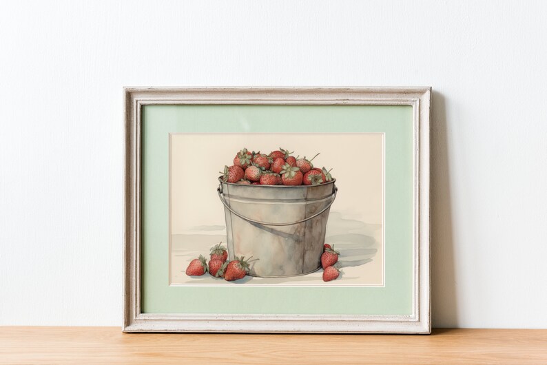 May include: A framed watercolor painting of a metal bucket filled with red strawberries. The bucket is sitting on a light brown surface. There are a few strawberries scattered around the bucket.