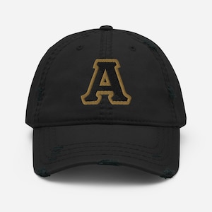 May include: Black distressed baseball cap featuring a gold-colored letter "A" on the front. The letter is outlined in a darker gold color. The cap has a curved brim and a distressed, worn look.