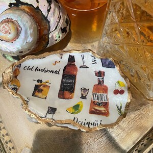 May include: A white ceramic coaster shaped like a seashell with a gold rim. The coaster has a colorful design of various cocktails and drinks, including "Old Fashioned", "Whiskey", "Tequila", and "Daiquiri".