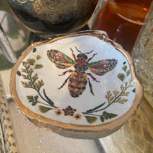 May include: A small white ceramic dish with a gold rim, featuring a colorful bee illustration and floral designs. The dish is perfect for holding jewelry or other small items.
