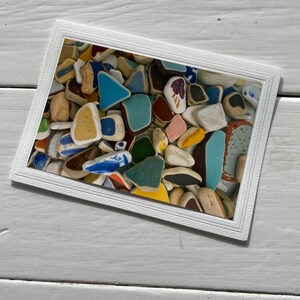 May include: A framed artwork featuring a collection of colorful sea glass pieces. The sea glass fragments are in various shapes and sizes, with colors including blue, green, yellow, and brown. The frame is white and rectangular, and the artwork is displayed on a white wooden surface.