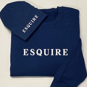 May include: A navy blue sweatshirt and beanie set. The sweatshirt and beanie both have the word "ESQUIRE" embroidered in white. The beanie is folded over and rests on the sweatshirt.