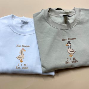 May include: Two light-coloured sweatshirts with embroidered geese. One sweatshirt is light blue with a goose wearing a flower crown and the text "His Goose J+M Est. 2025". The other sweatshirt is light green with a goose wearing a top hat and the text "Her Goose J+M Est. 2025".