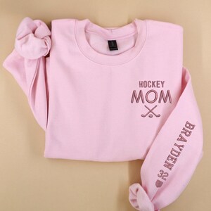 May include: A pale pink sweatshirt with the embroidered text "HOCKEY MOM" and crossed hockey sticks on the chest. The sleeve displays the name "BRAYDEN" and the number "32". The sweatshirt has a round neck and long sleeves.