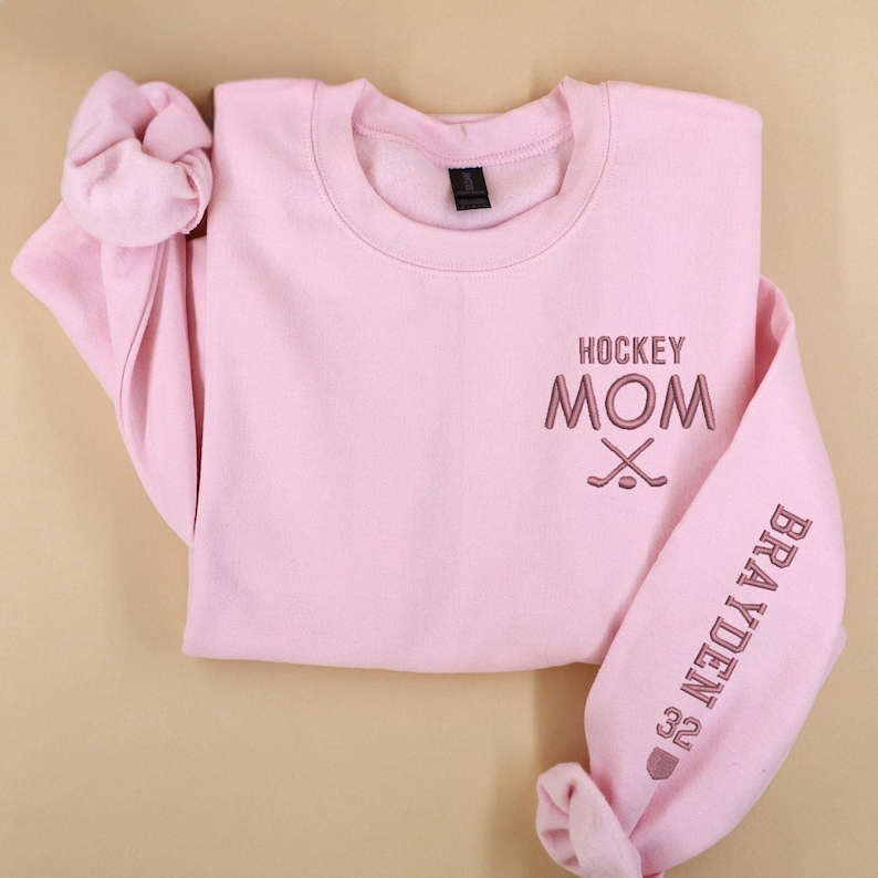May include: A pale pink sweatshirt with the embroidered text "HOCKEY MOM" and crossed hockey sticks on the chest. The sleeve displays the name "BRAYDEN" and the number "32". The sweatshirt has a round neck and long sleeves.