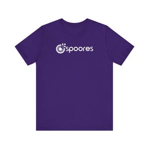 May include: A purple t-shirt featuring the word "Spoores" in white, with a stylized floral design above the text. The shirt has a crew neck and short sleeves, suitable for casual wear.