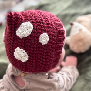May include: A burgundy crochet mushroom hat with white spots. The hat is handmade and appears to be for a baby or toddler. The hat is on a child wearing a light brown dress with mushroom graphics.