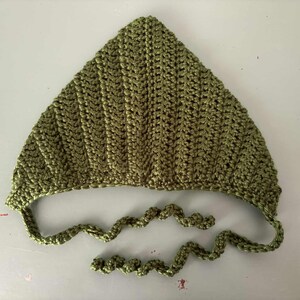 Pode incluir: Um gorro de croch&ecirc; verde-oliva feito &agrave; m&atilde;o com uma parte superior pontiaguda e la&ccedil;os presos. O gorro &eacute; feito com um ponto texturizado, e os la&ccedil;os s&atilde;o um cord&atilde;o torcido. O item &eacute; exibido em uma superf&iacute;cie branca.
