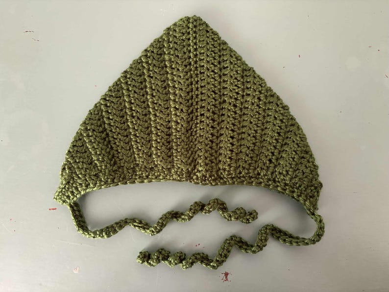 Pode incluir: Um gorro de croch&ecirc; verde-oliva feito &agrave; m&atilde;o com uma parte superior pontiaguda e la&ccedil;os presos. O gorro &eacute; feito com um ponto texturizado, e os la&ccedil;os s&atilde;o um cord&atilde;o torcido. O item &eacute; exibido em uma superf&iacute;cie branca.