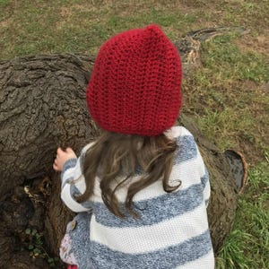 Pode incluir: Uma crian&ccedil;a usando um chap&eacute;u vermelho de croch&ecirc; e um su&eacute;ter listrado azul e branco. A crian&ccedil;a est&aacute; perto de um tronco de &aacute;rvore em uma superf&iacute;cie gramada. O chap&eacute;u &eacute; de cor vermelha escura s&oacute;lida.