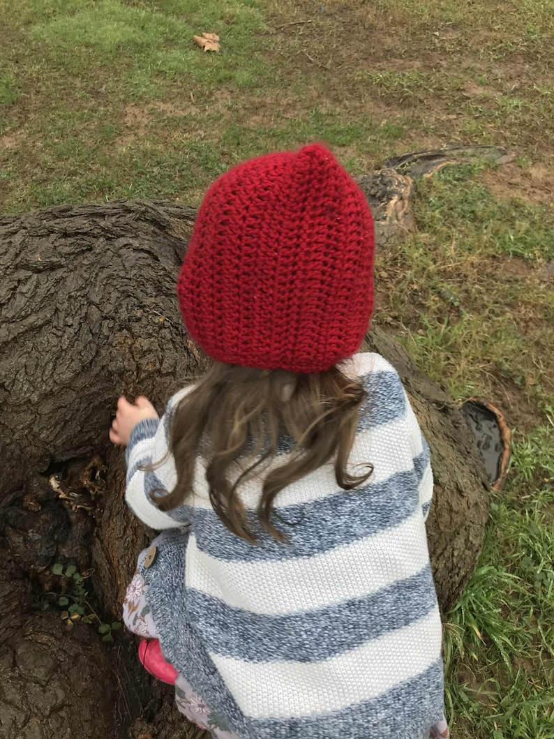 Pode incluir: Uma crian&ccedil;a usando um chap&eacute;u vermelho de croch&ecirc; e um su&eacute;ter listrado azul e branco. A crian&ccedil;a est&aacute; perto de um tronco de &aacute;rvore em uma superf&iacute;cie gramada. O chap&eacute;u &eacute; de cor vermelha escura s&oacute;lida.