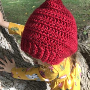 Pode incluir: Um chap&eacute;u de croch&ecirc; vermelho vibrante com uma ponta pontiaguda. O chap&eacute;u &eacute; feito de um fio texturizado e tem uma borda canelada. O chap&eacute;u &eacute; usado por uma pessoa que usa uma camisa amarela de manga comprida com um padr&atilde;o floral.