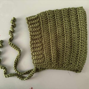 Pode incluir: Touca de croch&ecirc; verde-oliva com la&ccedil;os longos em espiral. A touca apresenta uma textura canelada e &eacute; feita de um fio macio. Este item feito &agrave; m&atilde;o &eacute; perfeito para beb&ecirc;s e crian&ccedil;as pequenas.