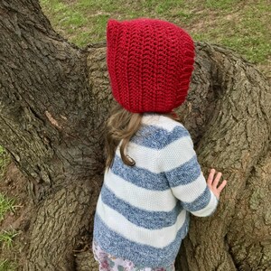 Pode incluir: Uma crian&ccedil;a vestindo um capuz vermelho brilhante de malha e um su&eacute;ter listrado azul e branco. A crian&ccedil;a est&aacute; perto de uma &aacute;rvore grande, com as m&atilde;os no tronco. O su&eacute;ter &eacute; combinado com uma saia estampada floral.