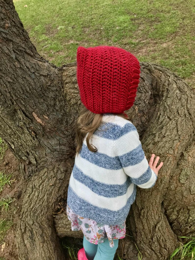 Pode incluir: Uma crian&ccedil;a vestindo um capuz vermelho brilhante de malha e um su&eacute;ter listrado azul e branco. A crian&ccedil;a est&aacute; perto de uma &aacute;rvore grande, com as m&atilde;os no tronco. O su&eacute;ter &eacute; combinado com uma saia estampada floral.