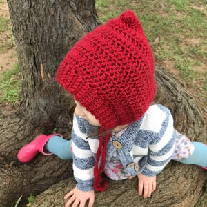 May include: A bright red, crocheted bonnet with long ties. The bonnet is worn by a child, paired with a blue and white striped cardigan and pink wellington boots. The bonnet is a vibrant shade of red.