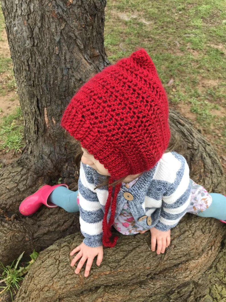 Pode incluir: Um gorro vermelho de croch&ecirc; feito &agrave; m&atilde;o com la&ccedil;os longos. O gorro &eacute; usado por uma crian&ccedil;a, combinado com um cardig&atilde; listrado azul e branco e botas rosa. O gorro &eacute; de um vermelho vibrante.