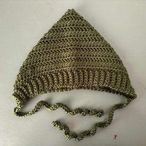 Pode incluir: Um chap&eacute;u de croch&ecirc; verde-oliva feito &agrave; m&atilde;o, com um topo pontudo e uma aba canelada. O chap&eacute;u tem la&ccedil;os longos e em espiral. O chap&eacute;u &eacute; feito de fio texturizado, adequado para beb&ecirc;s e crian&ccedil;as.