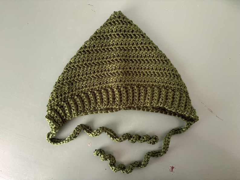 Pode incluir: Um chap&eacute;u de croch&ecirc; verde-oliva feito &agrave; m&atilde;o, com um topo pontudo e uma aba canelada. O chap&eacute;u tem la&ccedil;os longos e em espiral. O chap&eacute;u &eacute; feito de fio texturizado, adequado para beb&ecirc;s e crian&ccedil;as.