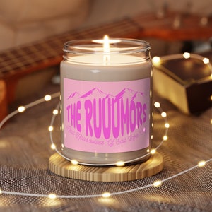 May include: A brown candle with a pink label that says "The Rumors Real Housewives of Salt Lake" with a graphic of mountains. The candle is on a wooden coaster.