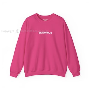 Op de afbeelding: Een felroze sweatshirt met lange mouwen en ronde hals, voorzien van het woord BRAVOHOLIC in witte hoofdletters in het midden van de borst. Het kledingstuk heeft een geribbelde halslijn, boorden en zoom met een eenvoudig ontwerp.
