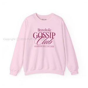 May include: A light pink crewneck sweatshirt featuring dark maroon text on the chest that reads Bravoholic GOSSIP Club with the subtext MEMBERS ONLY EST 2026. The garment has long sleeves, a ribbed neckline, and ribbed cuffs at the wrists and waist.