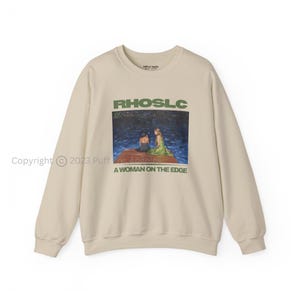 May include: A cream-coloured crew neck sweatshirt. The front displays a rectangular graphic with the text "RHOSLC" above an image of two women on a dock. Below the image is the text "A WOMAN ON THE EDGE."