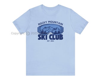 Könnte beinhalten: Hellblaues T-Shirt mit einem Grafikdesign. Das Design zeigt eine Berglandschaft in einer Skibrille, darüber der Text "Rocky Mountain Ski Club". Darunter steht "EST 2024".