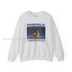 May include: A light gray crewneck sweatshirt featuring a graphic print on the chest. The print displays the text RHOSLC above an image of two figures on a wooden dock over dark water, with the text A WOMAN ON THE EDGE printed below the image.