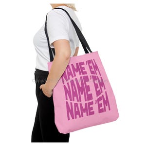 Puede incluir: Un bolso tote rosa con correas negras. El bolso presenta el texto "NAME 'EM" en un diseño curvo y repetitivo en un tono más oscuro de rosa. El bolso lo sostiene una persona.