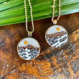 May include: Two necklaces with round pendants featuring a white background with brown mountains and a yellow sun. The pendants are on a wooden surface with green leaves.
