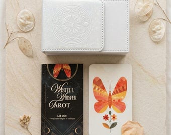May include: A white tarot card box with a circular embossed design, a black tarot deck box titled Wistful Whisper Tarot by Liz Ooi, and a single tarot card featuring an orange and yellow butterfly illustration above a small flower.