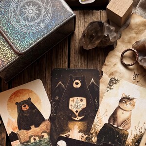 May include: A rectangular holographic storage box with a circular star design, three illustrated cards featuring a bear on a log, a bear with a geometric chest symbol, and a cat, a brown ring, and two raw crystal stones on a wooden surface.